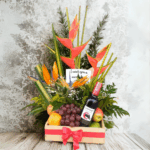 Arreglo floral tropical con heliconias y flores naranjas acompañado de uvas, peras, manzanas y una botella de vino tinto en caja de madera con lazo rojo