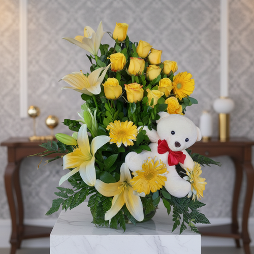 arreglo floral con rosas amarillas, gerberas y lirios blancos acompañado de un oso de peluche blanco con moño rojo sobre pedestal de mármol