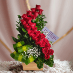 arreglo floral vertical con varias rosas rojas frescas, follaje verde y hojas anchas decorativas en base de madera clara sobre fondo difuminado rosa