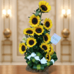 Arreglo vertical de girasoles amarillos con centro oscuro, hojas verdes y flores blancas en la base, colocado sobre una mesa de madera con fondo interior desenfocado