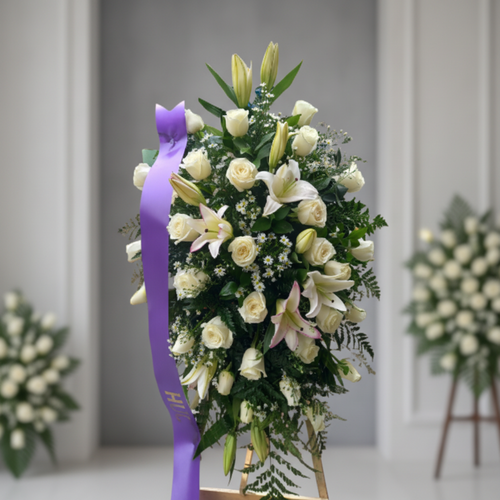 Arreglo floral funerario en forma de corona con rosas y lirios blancos, decorado con cinta morada, sobre caballete en interior elegante