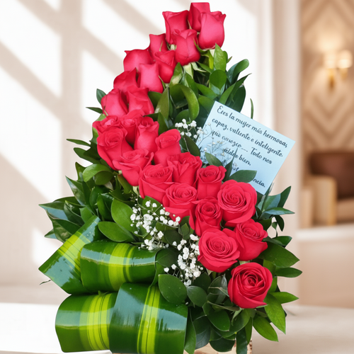 Arreglo floral de rosas rojas con tarjeta dedicatoria Arreglo de rosas rojas en forma escalonada con follaje verde, lazos de hojas y una tarjeta con mensaje en español sobre fondo interior desenfocado