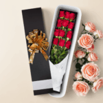 Caja de regalo alargada negra con lazo dorado que contiene un ramo de rosas rojas envueltas en papel blanco, junto a rosas rosadas sobre fondo beige