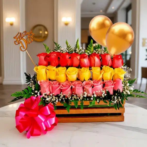Arreglo floral Escala con rosas rojas, amarillas y rosadas en caja de madera, con globos dorados y lazo rosa, sobre mesa de m