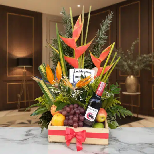 Arreglo floral Platonia con heliconias, aves del paraíso, uvas, manzanas, duraznos y botella de vino tinto, sobre mármol en u