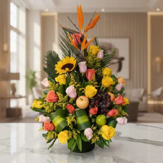 Exquisito Arreglo Floral con Frutas Tropicales para Decoración Elegante Arreglo floral vibrante con rosas amarillas y salmón, girasol, ave del paraíso y frutas tropicales como uvas, manzana y lima,