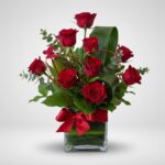 Arreglo floral ROSETTA con seis rosas rojas frescas, follaje verde y un lazo rojo en un jarrón de cristal, ideal para regalar