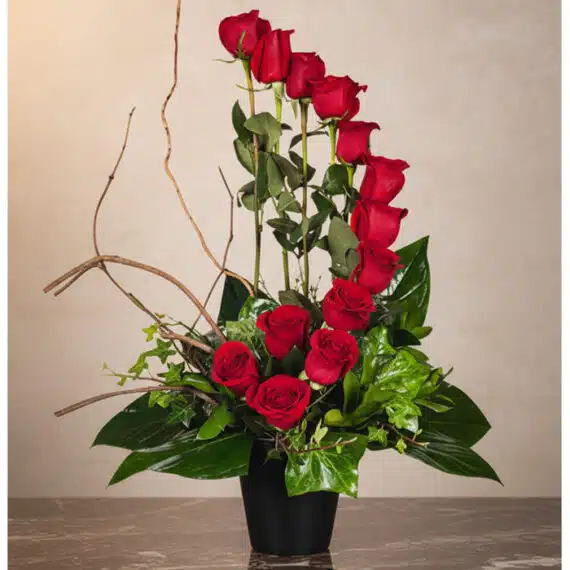 Arreglo GIANNA: 13 Rosas Rojas, Símbolo de Amor y Pasión Arreglo floral GIANNA con 13 rosas rojas frescas en un jarrón negro, ramas decorativas y follaje verde, sobre una superficie