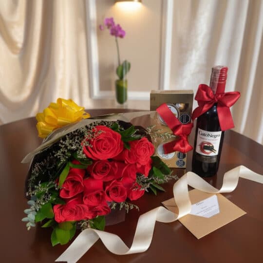 Elegante bouquet de rosas rojas, botella de vino tinto Gato Negro y caja de chocolates Ferrero Rocher sobre mesa de madera pu