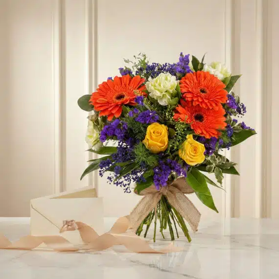 Bouquet Floral SABRINA con rosas amarillas, gerberas naranjas, flores moradas y follaje verde, atado con cinta de yute, sobre