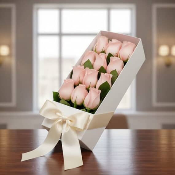 Caja de 12 Rosas Rosadas: El Regalo Perfecto para Cualquier Ocasión Caja blanca inclinada con 12 rosas rosadas frescas y un moño de satén, sobre una mesa de madera oscura con un fondo elegante