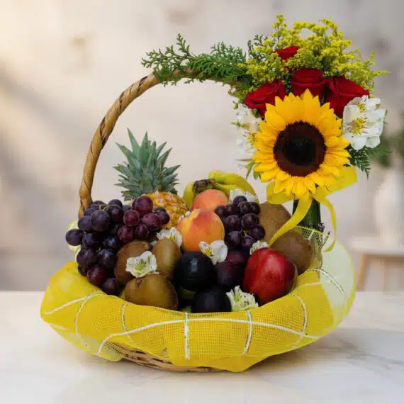 Canasta de Frutas Draco con uvas, manzanas, duraznos, kiwis, piña y arreglo floral de rosas, girasol y flores blancas sobre m