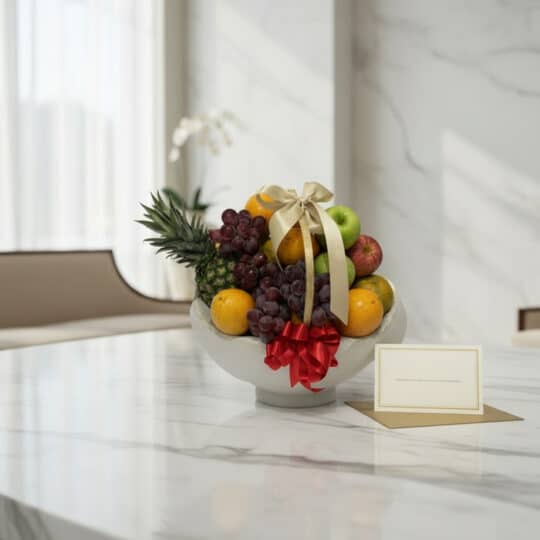 Canasta de frutas gourmet con piña, uvas, manzanas y naranjas, decorada con cintas doradas y rojas, sobre una mesa de mármol