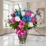 Jarrón Primavera con un arreglo floral vibrante de rosas blancas, gerberas fucsia y alstroemerias azules, sobre una superfici