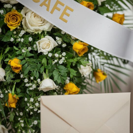 Arreglo floral fúnebre con rosas blancas y amarillas, follaje verde, rocío sutil y cinta satinada. Detalle de sobre de condol