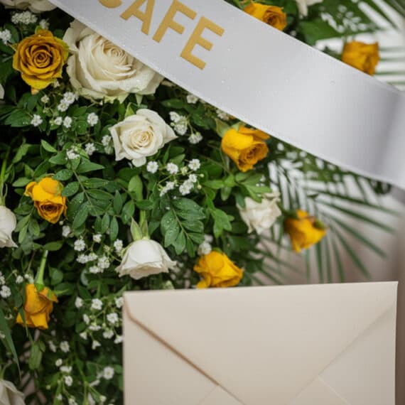 Arreglo floral fúnebre con rosas blancas y amarillas, follaje verde, rocío sutil y cinta satinada. Detalle de sobre de condol